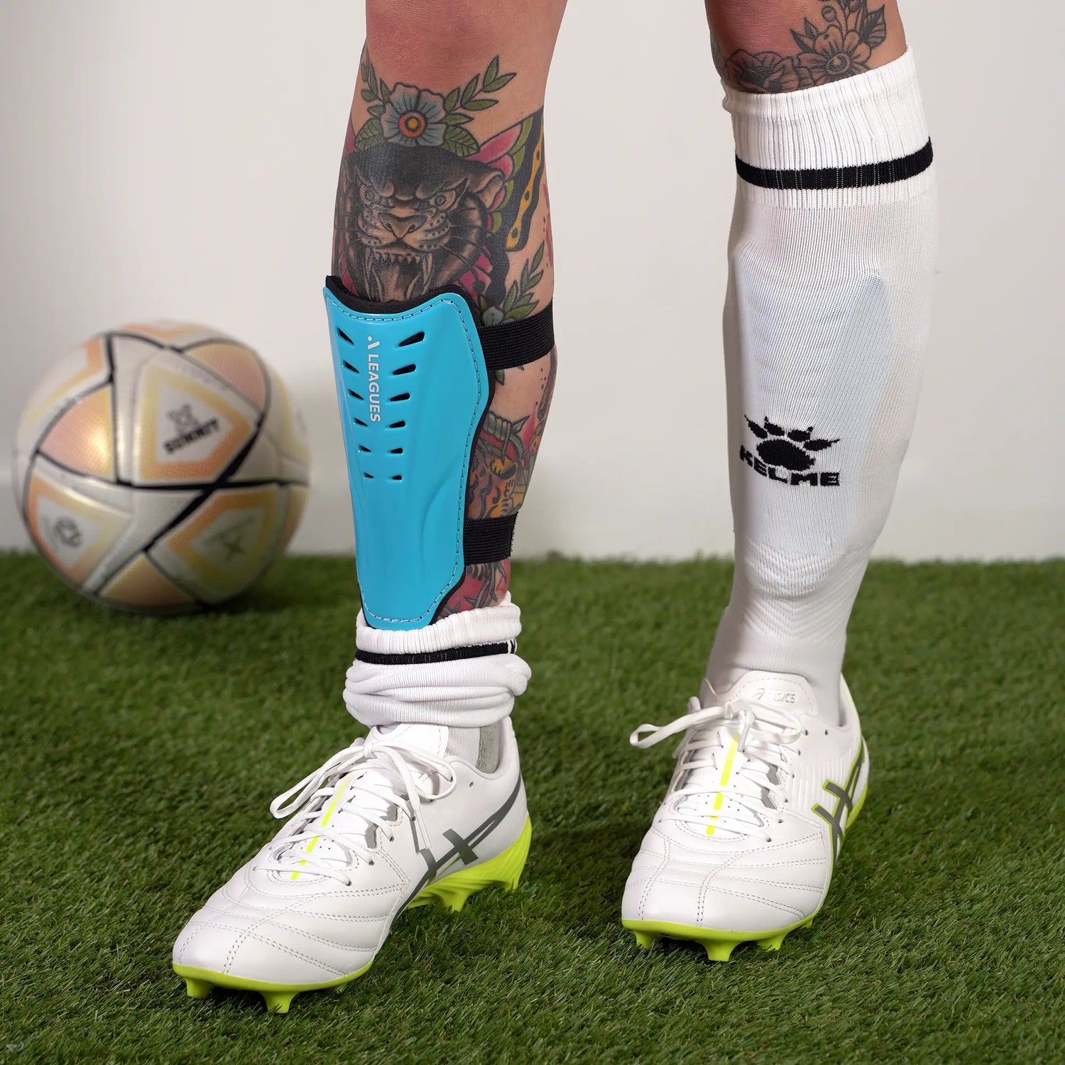 Person wearing white soccer cleats with a blue shin guard on grass, with a soccer ball in the background.