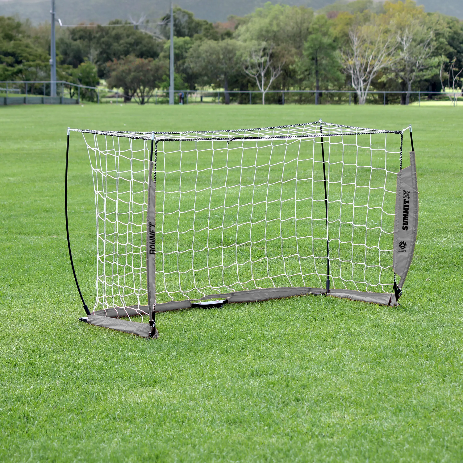 Bownet 1x2m portable soccer goal with white netting and gray base pads, set up on a green grass field with trees in the background. Football Australia approved 100cm x 120cm goal by SUMMIT Sport.