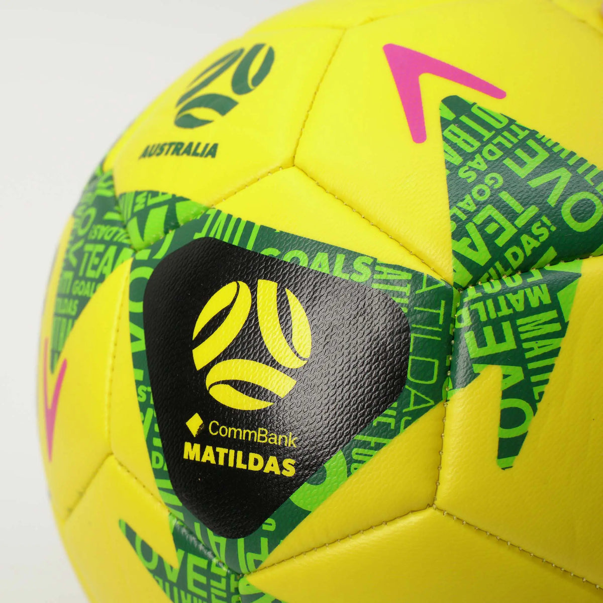Close-up of a yellow soccer ball with green and black patterned panels, a black CommBank Matildas logo patch, and pink arrow accents on a light gray background.