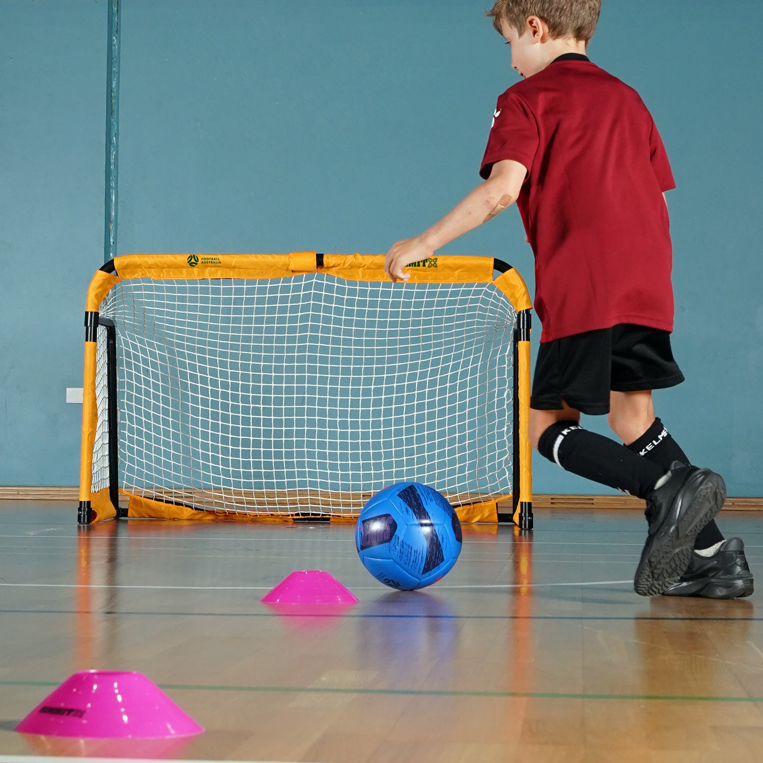 Child playing with a blue ball near a small soccer goal on a gymnasium floor.  Portable soccer goal that folds for easy transport. Use indoors or outdoors for kids or soccer players. 