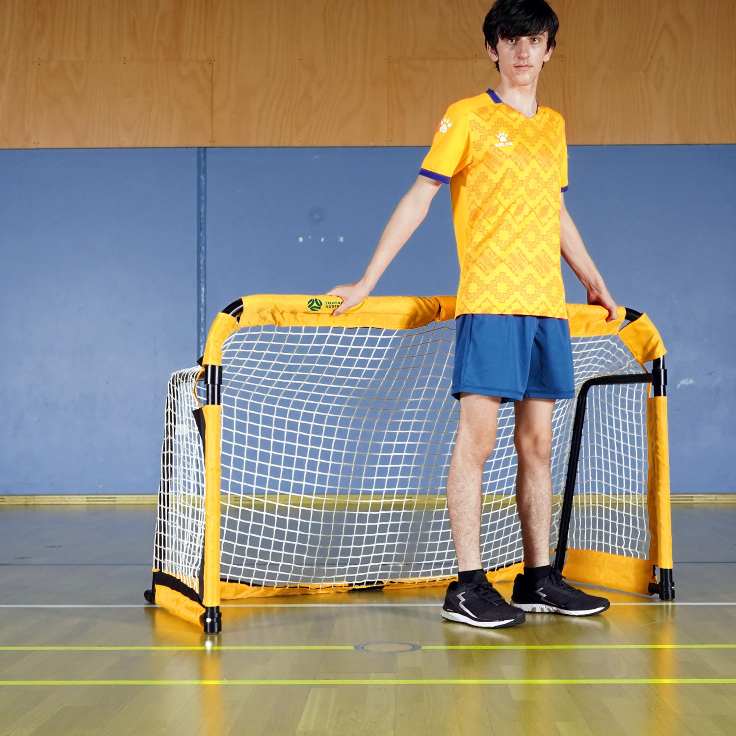 Aluminium portable style soccer goal 5ft size. Person in a yellow sports jersey and blue shorts standing next to a small soccer goal on a court.