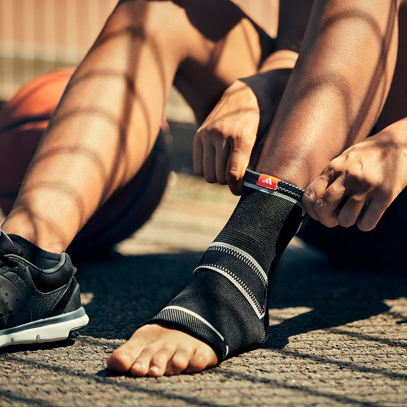 Person pulling an adidas ankle support over their foot, with a basketball in background. 