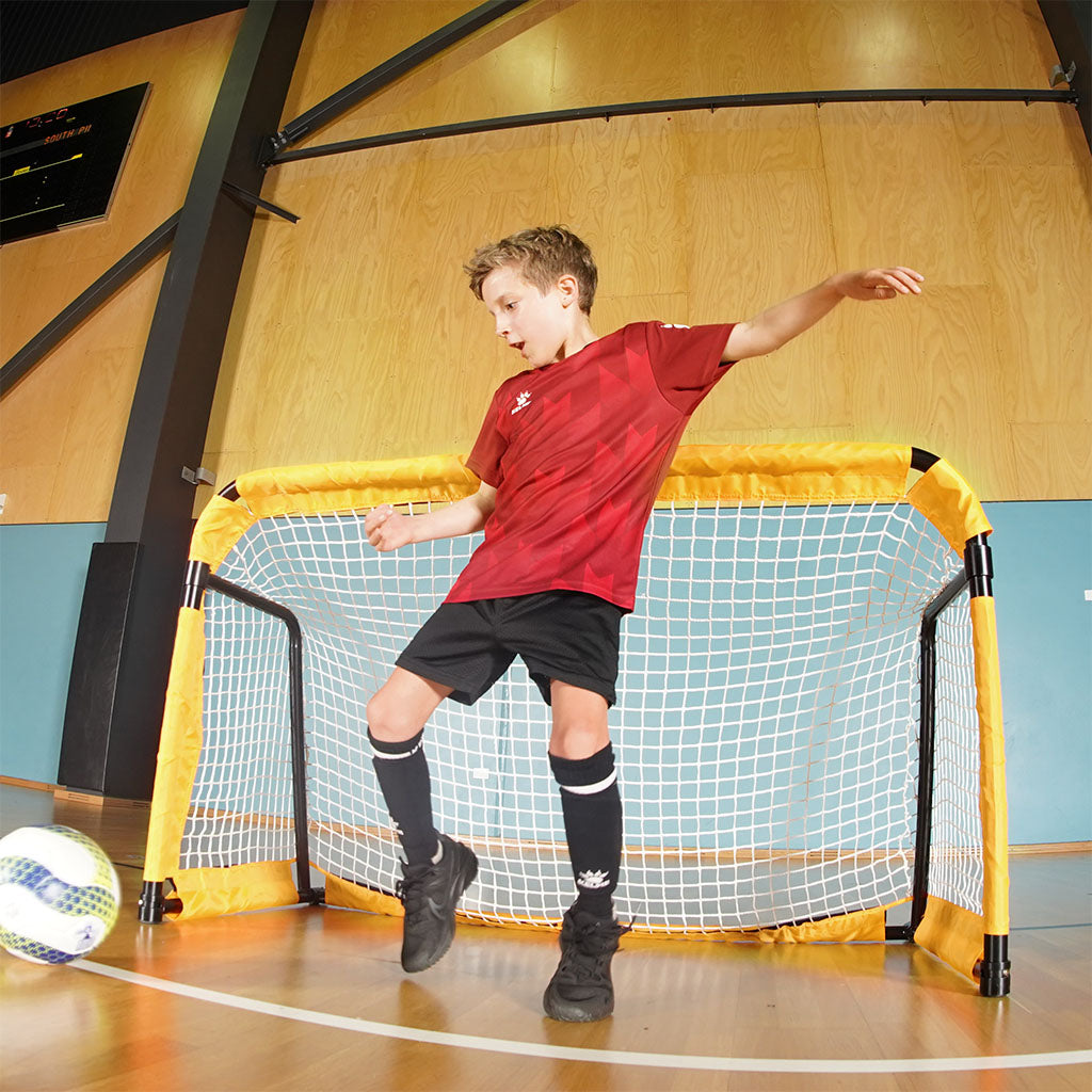 Child playing with a futsal ball in front of a SUMMIT Football Australia metal folding goal. Aluminium style soccer goal 6ft in size indoors with kid enjoying playing a game of soccer.