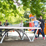 CORNILLEAU Pro Park Outdoor Table Cornilleau being used by kids in a park outside in a public space. Kids are having a game of ping pong, with young girl hitting the ball over the table tennis net.