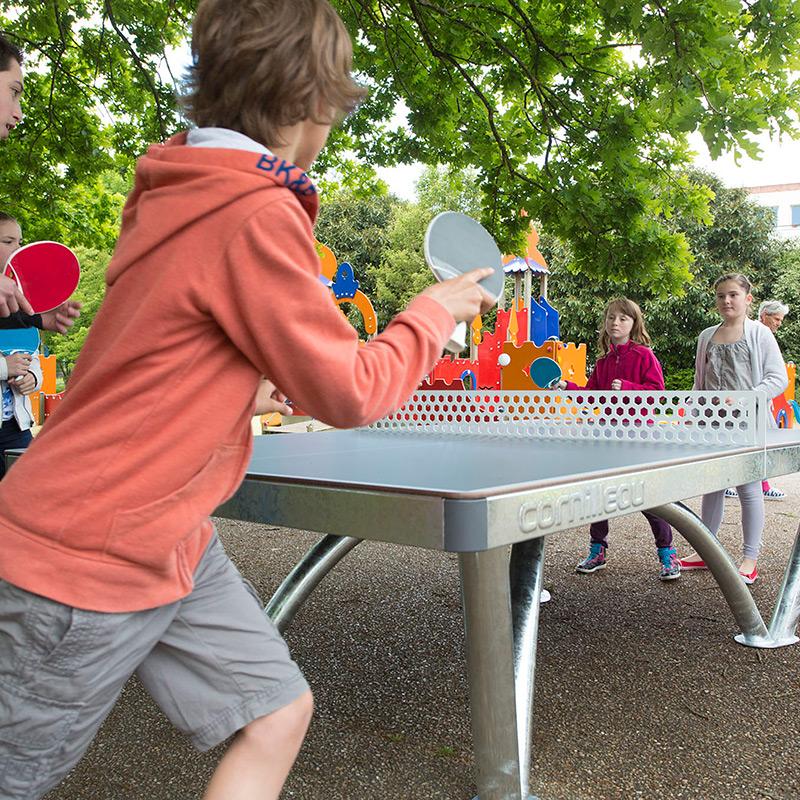 CORNILLEAU Pro Park Outdoor Table Cornilleau close up in the middle of a public game. Young boy is kitting the ball over the ping pong net towards a young girl holding a ping pong bat.