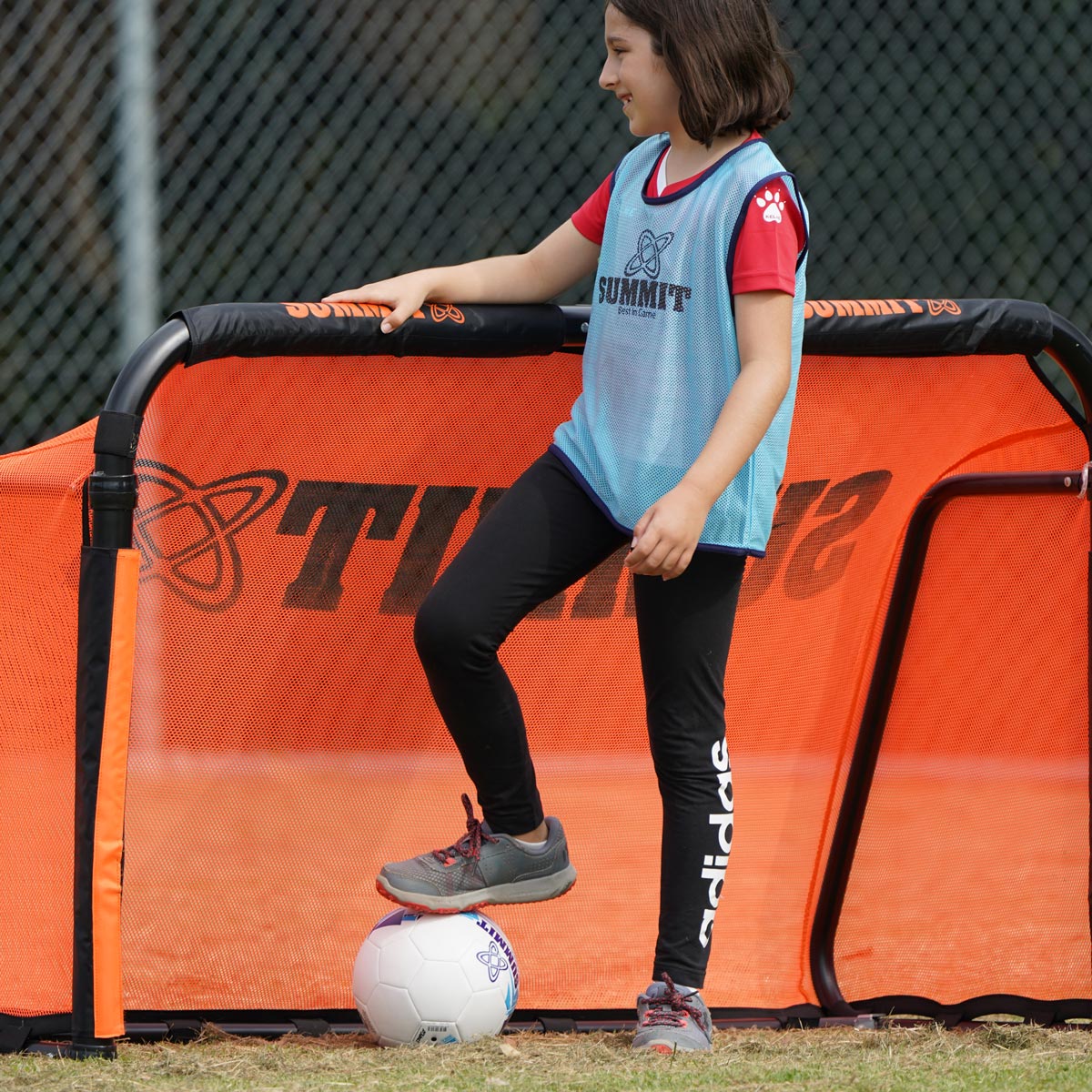 Young girl playing soccer in blue sports bib, size 4 soccer ball and orange football goal.